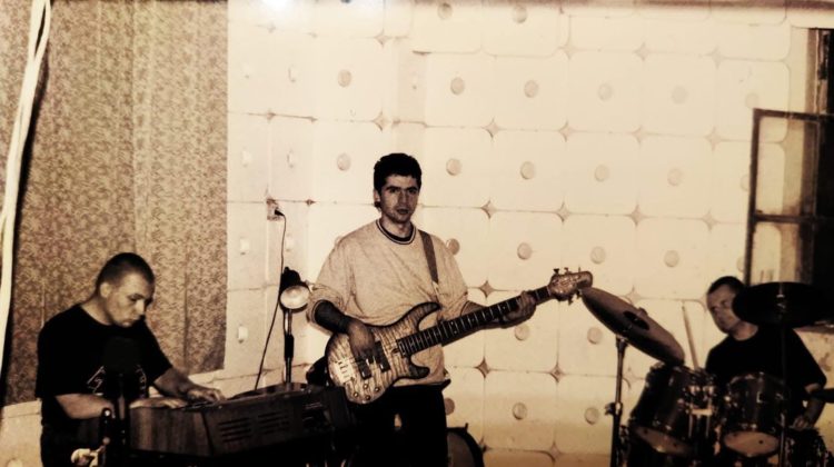 Czarno-białe zdjęcie przedstawia trzech muzyków podczas próby lub występu w pomieszczeniu o wyciszonych ścianach. Po lewej stronie mężczyzna gra na keyboardzie. W centrum kadru znajduje się mężczyzna z gitarą basową, patrzy w stronę aparatu. Po prawej stronie za perkusją (z widocznym napisem Yamaha) siedzi trzeci muzyk. Całość utrzymana jest w klimacie retro, przypominającym lata 80. lub 90. Pomieszczenie wygląda na salę prób lub studio nagraniowe, a ściany pokryte są materiałem dźwiękochłonnym.