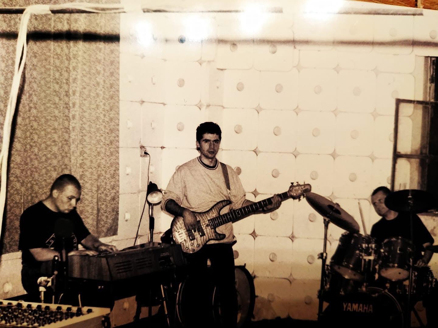Czarno-białe zdjęcie przedstawia trzech muzyków podczas próby lub występu w pomieszczeniu o wyciszonych ścianach. Po lewej stronie mężczyzna gra na keyboardzie. W centrum kadru znajduje się mężczyzna z gitarą basową, patrzy w stronę aparatu. Po prawej stronie za perkusją (z widocznym napisem Yamaha) siedzi trzeci muzyk. Całość utrzymana jest w klimacie retro, przypominającym lata 80. lub 90. Pomieszczenie wygląda na salę prób lub studio nagraniowe, a ściany pokryte są materiałem dźwiękochłonnym.