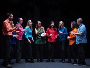 obraz grupa dziewięciu osób stojących w półokręgu na scenie, ubranych w kolorowe koszule i bluzki: czerwone, pomarańczowe, zielone, niebieskie, różowe i błękitne. Każda osoba wydaje się być zaangażowana w śpiewanie lub rozmowę, utrzymując kontakt wzrokowy ze sobą nawzajem. Jedna kobieta, stojąca w środku, trzyma instrument perkusyjny typu bębenek. W tle ciemne, jednolite tło, co podkreśla sceniczny charakter zdjęcia. Wszyscy uczestnicy mają uśmiechnięte twarze i wyglądają na rozluźnionych oraz radosnych