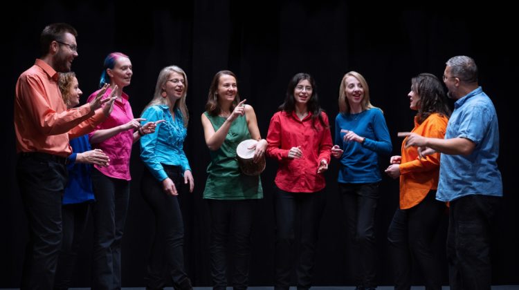 obraz grupa dziewięciu osób stojących w półokręgu na scenie, ubranych w kolorowe koszule i bluzki: czerwone, pomarańczowe, zielone, niebieskie, różowe i błękitne. Każda osoba wydaje się być zaangażowana w śpiewanie lub rozmowę, utrzymując kontakt wzrokowy ze sobą nawzajem. Jedna kobieta, stojąca w środku, trzyma instrument perkusyjny typu bębenek. W tle ciemne, jednolite tło, co podkreśla sceniczny charakter zdjęcia. Wszyscy uczestnicy mają uśmiechnięte twarze i wyglądają na rozluźnionych oraz radosnych